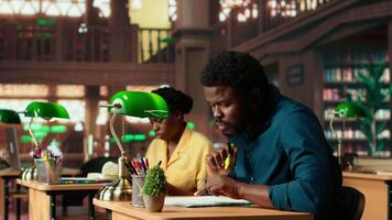 African american pupil underlining key words and phrases at the library, creating a summary with the highlighter pen at a university campus study space. Academic research for exams. Camera B. video