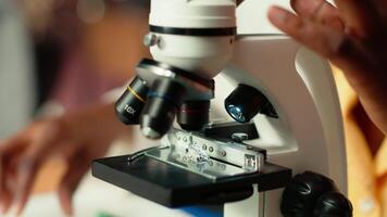 Future researcher works on her thesis by examining microorganisms under the microscope lenses, reading about microbiology and medical science. Focus on scientific exams. Close up. Camera B. video