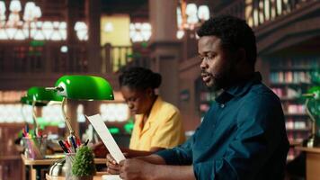 University student researches academic topics in a traditional library, using the classic study space to review lessons and organize course materials. Completing class notes for exams. Camera B. video