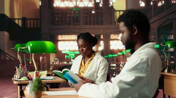 Young medical students comparing knowledge and class notes at the library, studying together to obtain their residency. Pupils learning healthcare methods by reading surgery notes. Camera B. video