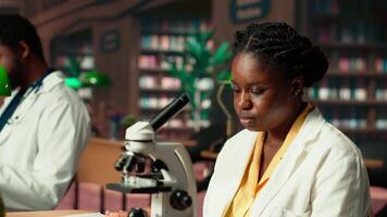 Black student researching biotechnology in an university library, reviewing cells and microorganisms on a microscope. Learning for a successful thesis project that guarantees her career. Camera B. video