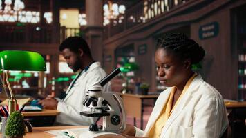 Black student researching biotechnology in an university library, reviewing cells and microorganisms on a microscope. Learning for a successful thesis project that guarantees her career. Camera A. video