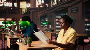 Focused african american student sits in a quiet library reading papers and class notes, studying under the warm glow of a green lamp. Woman surrounded by books and learning tools. Camera A. video
