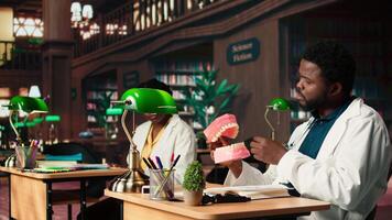 Medical student practices dentistry techniques with a plastic jaw model in a classic library, doing physical research for stomatology thesis. Developing professional skills for the career. Camera A. video