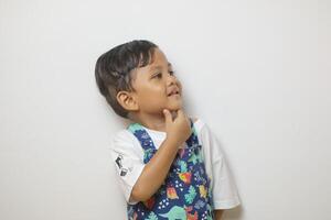Thoughtful young boy with curly hair looking aside at copy space, touching chin in contemplation, wearing blue shirt on yellow background, embodying curiosity and deep thought photo