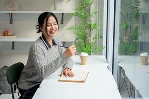 A positive Asian woman sits at a table by the window with her diary placed on the table. photo