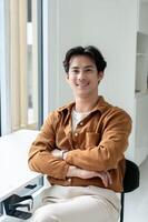 Handsome, positive Asian man sits at a table with his arms crossed in a room, smiling at the camera. photo