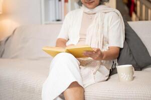 A close-up of a happy Asian retired senior woman relaxes on a sofa, enjoying reading a book. photo