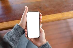 A top view of a woman holds a smartphone with a white screen mockup while sitting at a table indoors photo