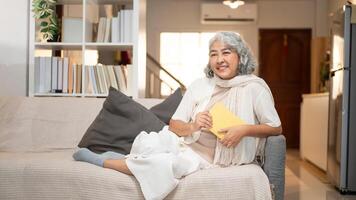 A happy Asian retired woman relaxes on a sofa, contemplating about happy life while reading a book. photo
