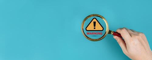 Danger and error alert concept. Hand uses magnifying glass to find symbol notification warning triangle on blue background, with copy space. Maintenance of security systems, and antivirus protection photo