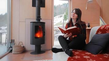 A woman enjoys reading a book while sitting by a warm fireplace in a modern wooden cabin, Cozy Winter Reading by the Fireplace in Cabin, video