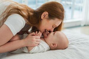 Happy family at home. Mother playing with little toddler child on bed. Mom baby relax playing having fun together. Mother looks at baby with love care. Mom of breast feeding baby rest in living room photo