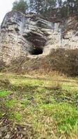 The tall rock face features a dark cave entrance framed by vibrant grass, highlighting the contrast between natures beauty and the stones rugged texture, inviting exploration video