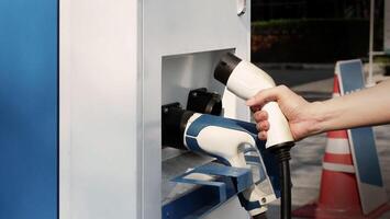 Driver of electric car hold batterie charger plug. Hand holding EV charging plug in front of camera with blurred background of outdoor. Sustainable energy powered electric charging station background photo