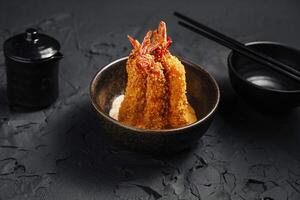 Crispy breaded shrimp served in a dark bowl with condiments and chopsticks on a textured black surface photo