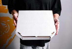 Individual holding a blank white pizza box in a casual setting with a colorful wall background photo