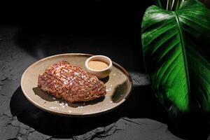 Perfectly seared steak served on a textured plate with a small bowl of sauce and a tropical leaf accent photo