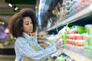 young African American woman in a work apron and disposable gloves works in a supermarket, stands among the shelves and lays out products and merchandise. photo