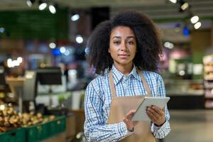 Portrait of young smiling African American woman, supermarket owner and worker standing in grocery department in apron, holding and using tablet, looking at camera. photo