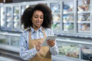A young smiling African-American woman works in a supermarket, in a work uniform, standing on the background of the dairy department and using a tablet. photo