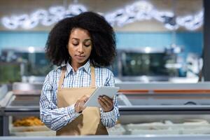 Worker in apron using tablet at fish department in supermarket. Image shows young woman checking inventory with modern technology. Concept of retail, seafood, fish, and grocery store. photo
