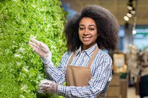 Supermarket worker wearing gloves and apron, and arranging fresh greens in the grocery store. Smiling, professional service, and healthy food concept. photo