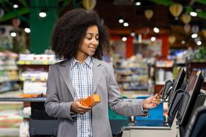 A smiling woman in a grey blazer uses a self-checkout kiosk at a grocery store. She holds an orange product while interacting with the machine. photo
