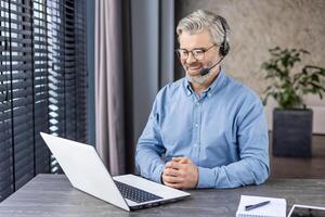 Satisfied senior entrepreneur in headset with microphone looking at portable computer screen with hands folded on desk. Pleased boss listening to reports of employees with improved results at home. photo