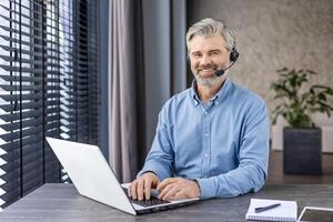 Senior grey-haired man in wireless headset sitting in home office with laptop and notebook. Smiling entrepreneur getting ready for online conference and discussing planning of working strategy. photo