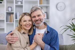 A cheerful middle-aged couple hugs warmly in their stylish living room, smiling at the camera with a bookshelf and modern decor in the background. photo