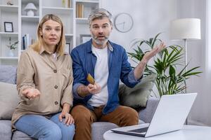 Middle-aged couple sitting on couch looking confused, holding a credit card, with laptop in front of them. Discussing uncertain financial issue or online payment problem. photo