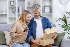 A cheerful mature couple sitting on a couch at home as they open a package together, showing excitement and curiosity. They are looking into the box with happy expressions. photo
