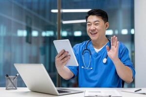 Asian man doctor in blue scrubs engaging in call on tablet, smiling and waving, showcasing communication and technology in healthcare, laptop and phone visible on desk. photo