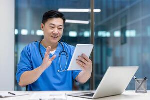 Asian man doctor in blue scrubs using tablet for virtual consultation in office. Engaged in call, smiling confidently, with laptop on desk, conveying professionalism and technology. photo