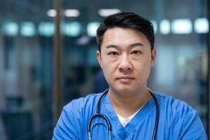 Asian man doctor in blue scrubs stands confidently with stethoscope in modern healthcare facility. Expression serious, projecting professionalism and dedication in medical field. photo