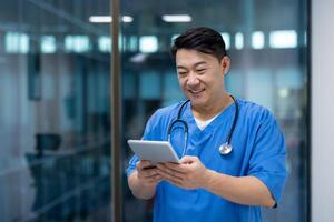 Asian man doctor in scrubs using tablet computer, smiling while checking medical records. Technology in healthcare enhances patient care, facilitating communication within medical team. photo