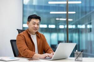 Asian man businessman using laptop in office setting. Engaged in work tasks, showcasing focus and productivity. Modern workspace reflects professional environment and technology use. photo