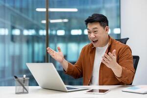 Asian man businessman with bright smile in modern office using laptop, expressing excitement and success during call. Achieving goals, enhancing productivity with innovative collaboration tools. photo