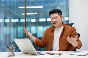 Asian man businessman showing frustration while working on laptop in office. Expressing stress and confusion with hands up and bewildered expression, phone on desk. photo