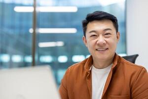 Asian man businessman smiling confidently in modern office environment with laptop. Bright professional setting with large windows, casual attire emphasizing relaxed workspace and productivity. photo