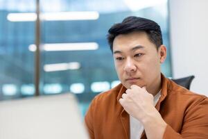 Asian man businessman in office working thoughtfully on laptop, concentrating on tasks and ideas in modern setting, showcasing business and technology concepts photo