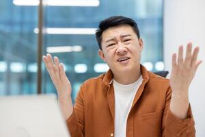 Asian man showing confusion in office, wearing casual brown jacket, sitting at desk with raised hands and puzzled expression. Office setting suggests business context and workplace scenario. photo