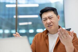 Asian man businessman showing frustrated expression at work with laptop in modern office. Scene of confusion or misunderstanding, highlighting workplace stress or problem-solving. photo