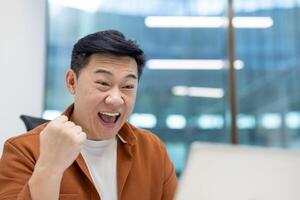 Asian man businessman showing excitement celebrating success at work with fist pump. Displaying positive energy in modern office, using laptop, showcasing motivation and achievement. photo