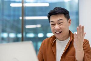 Asian man businessman in modern office having call on laptop, expressing excitement and enthusiasm. Engaged in virtual meeting showcasing technology use in workspace. photo