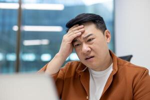 Asian man businessman feeling stressed at work while concentrating on laptop. Office environment with technology and challenges. Business setting showing pressure and problem-solving mindset. photo