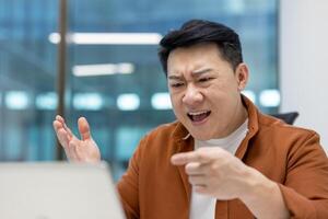 Asian man showing anger during call in office environment. Business professional using laptop communicates frustration, pointing and gesturing. Concept of workplace stress and remote meetings. photo