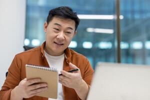 Asian man businessman engaging in virtual meeting. Holding notebook and pen while discussing professional topics. Demonstrates focus on collaboration and effective communication in modern office. photo