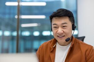 Asian man businessman with headset smiling while engaged in virtual communication. Captured in modern office environment showing professional interaction via technology. photo